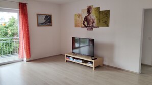 A living room with a flat-screen TV on a wooden stand, a painting on the wall, and a window with a red curtain. - Rottenburg City 4 rooms with cathedral view (Rottenburg)