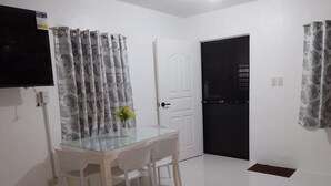 Een eethoek met een glazen tafel en witte stoelen, een tv aan de muur en kamerhoge gordijnen met een patroon.