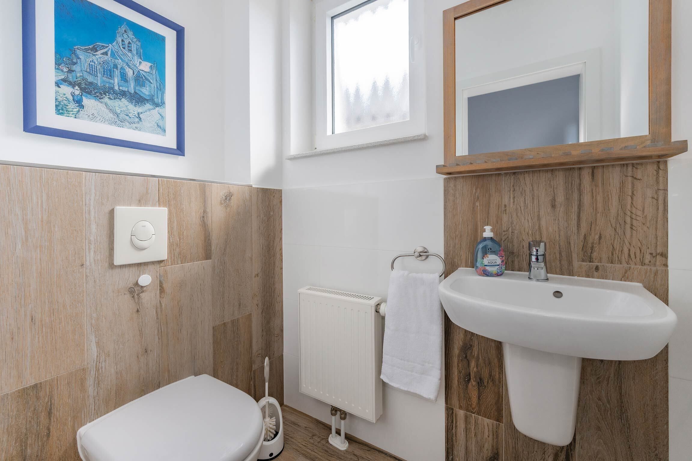 A bathroom with a white toilet, a white sink, a towel hanging on a rack, a radiator, and a framed painting on the wall.