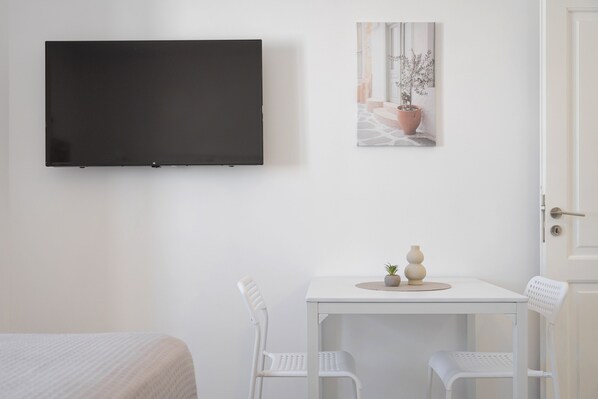 A room with a bed, a white table with two chairs, a flat-screen TV mounted on the wall, and a framed picture above the table. - Palma Elegant Suites (Naousa)