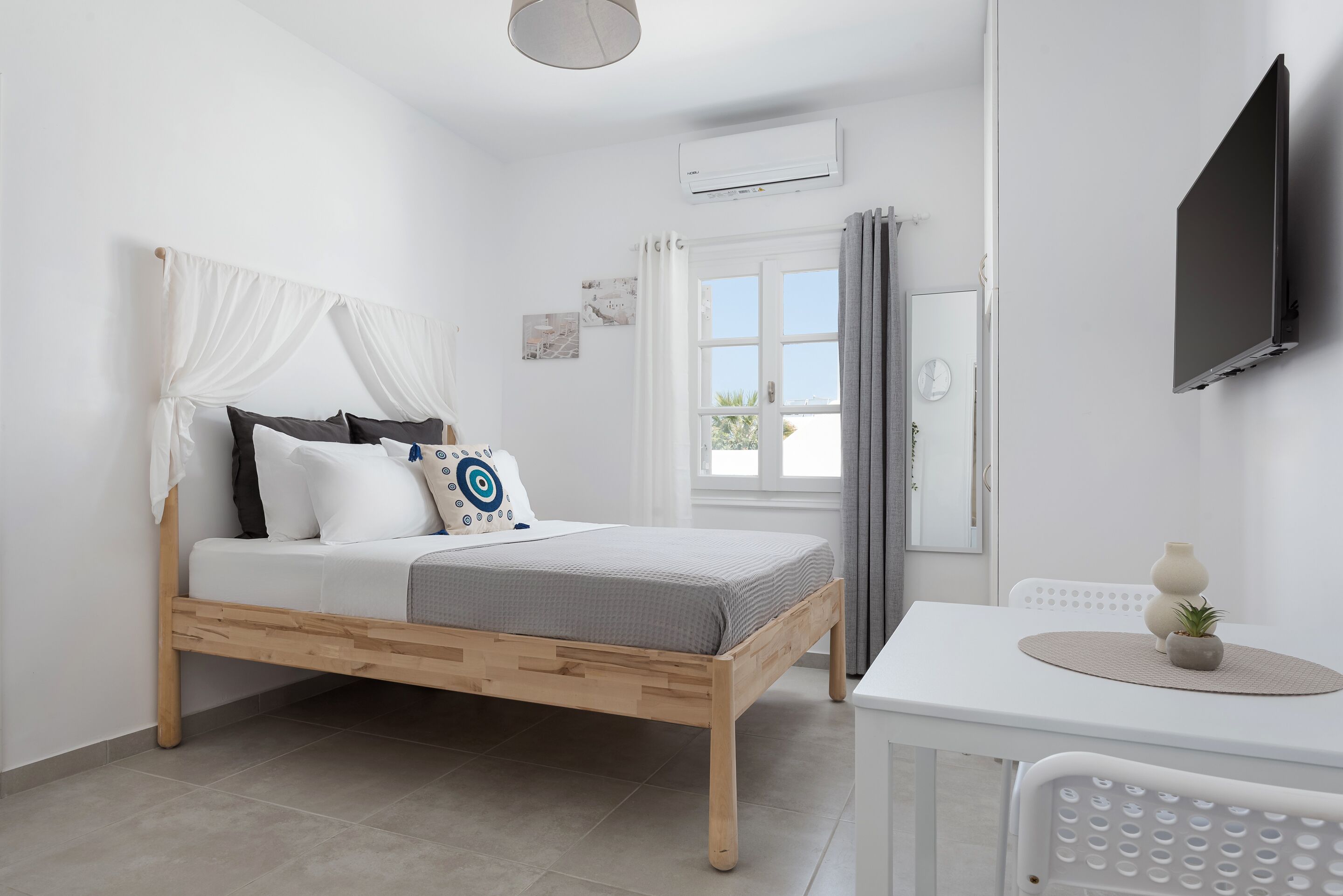 A bedroom with a wooden bed, white walls, a window with curtains, a white table with a vase, and a mounted TV.