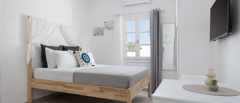 A bedroom with a wooden bed, white walls, a window with curtains, a white table with a vase, and a mounted TV.