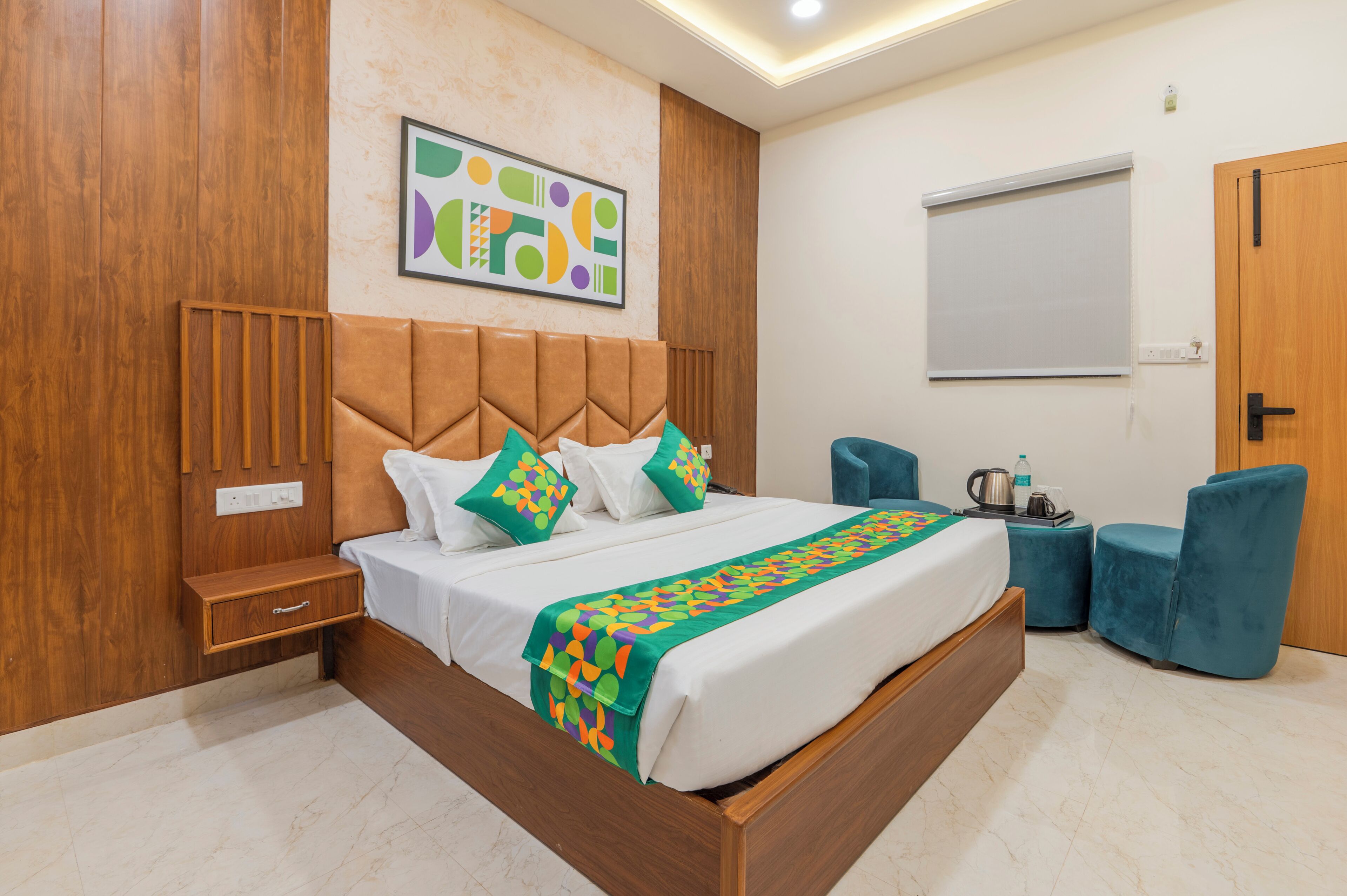 A modern hotel room with a wooden headboard, a large bed with a colorful blanket, a side table, a blue chair, and a whiteboard on the wall.
