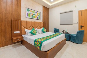 A modern hotel room with a wooden headboard, a large bed with a colorful blanket, a side table, a blue chair, and a whiteboard on the wall.