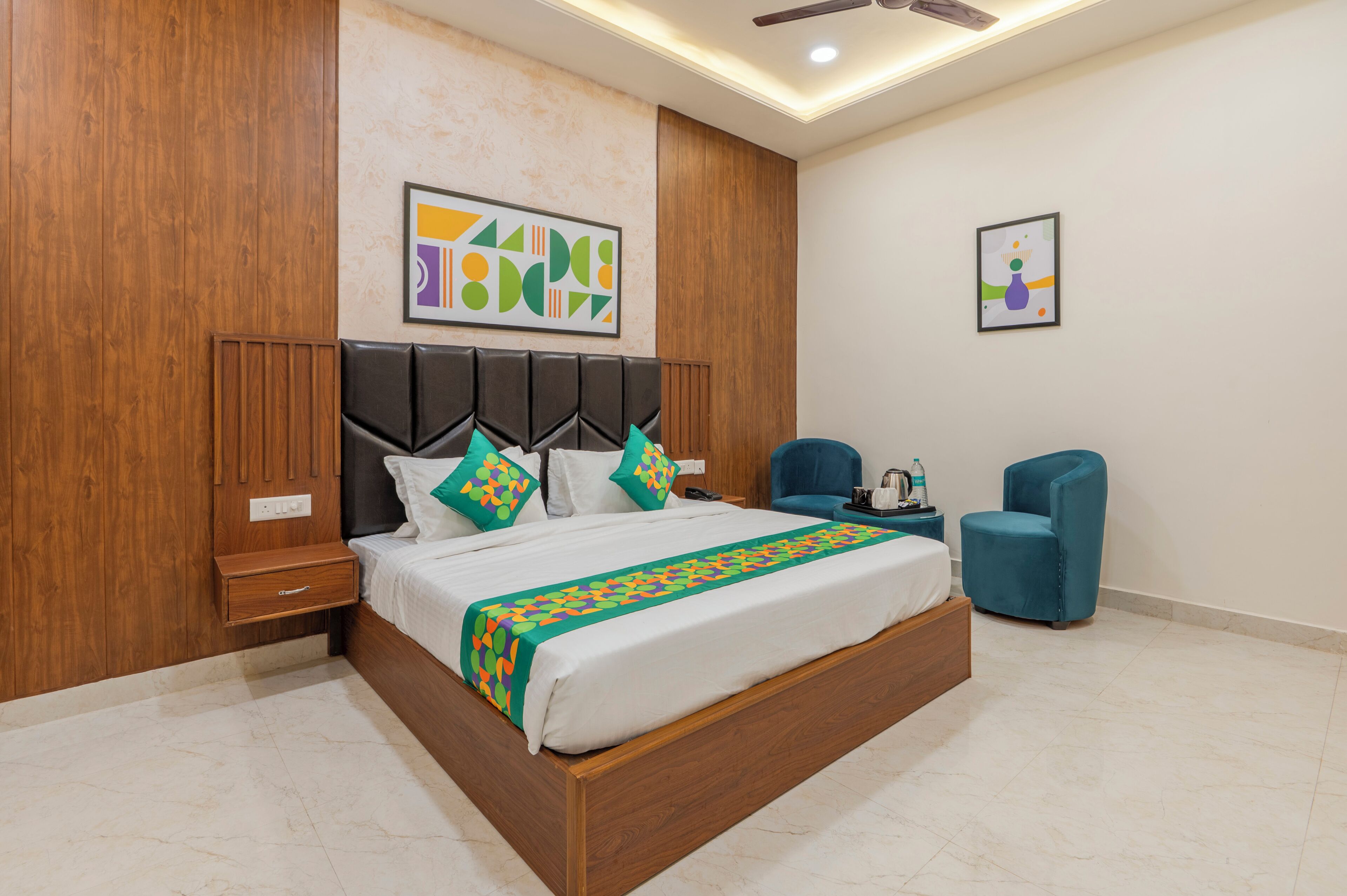 A modern bedroom with a wooden headboard, a bed with a colorful bedspread, two blue armchairs, and artwork on the walls.