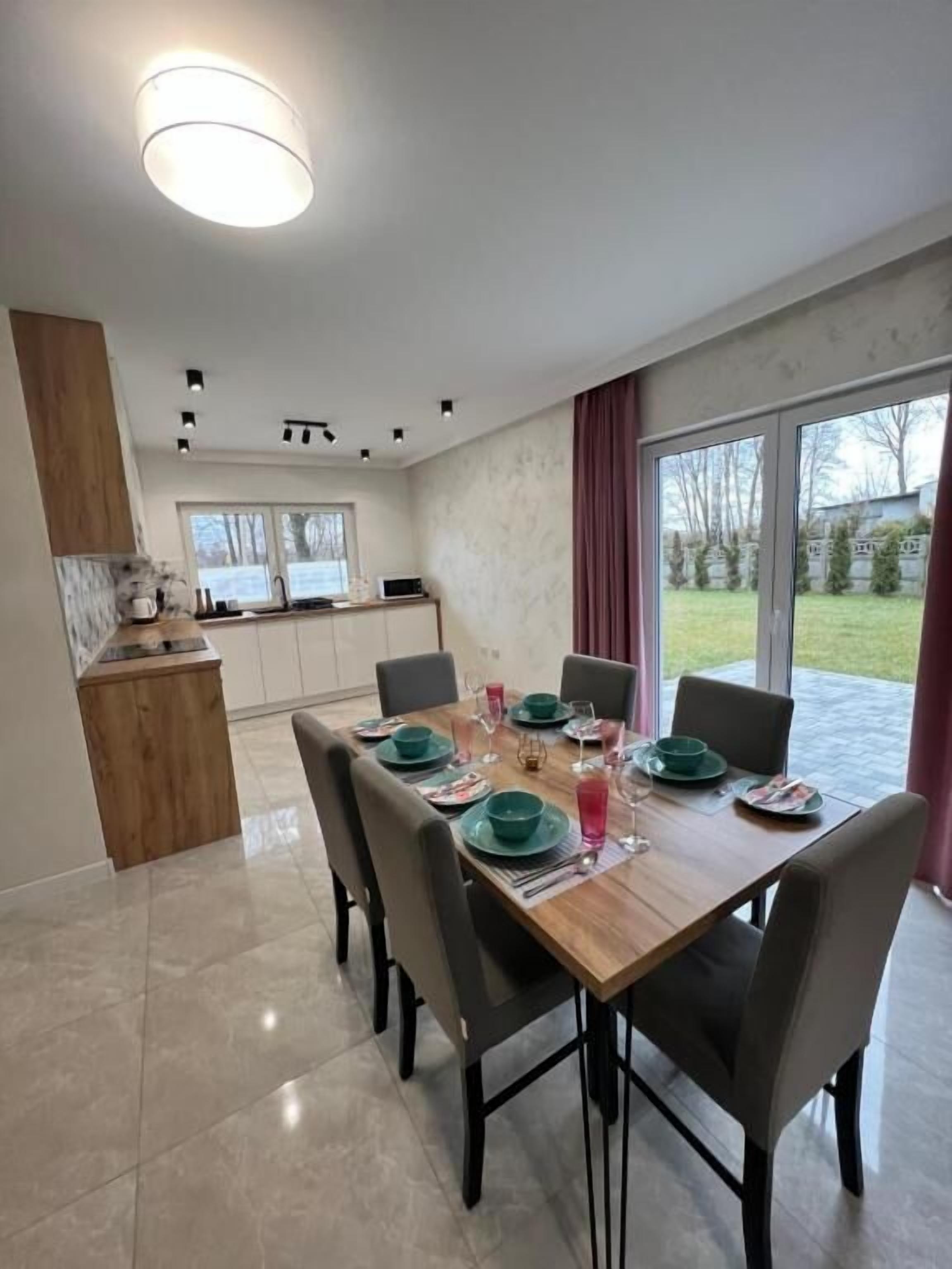A dining area with a wooden table set for four, a kitchen in the background, and a view of the outdoors.