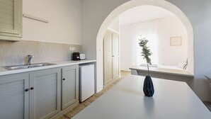 A modern kitchen with a white countertop, a built-in oven, and a coffee machine. There is a bench under an archway leading to a living area with a plant and a framed picture on the wall. - Phaedrus Living: Paros Cycladic Residences 8 (Paros)