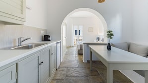 A modern kitchen with white cabinetry, a stainless steel sink, and a coffee machine. There is a stone floor leading to a dining area with a white table and a plant. An arched doorway leads to a balcony with a view. - Phaedrus Living: Paros Cycladic Residences 8 (Paros)
