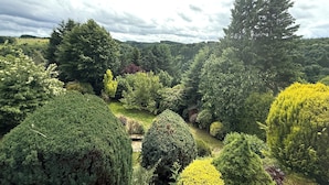 A lush garden with various trees and shrubs, a well-maintained lawn, and a distant view of a hilly landscape under a cloudy sky.