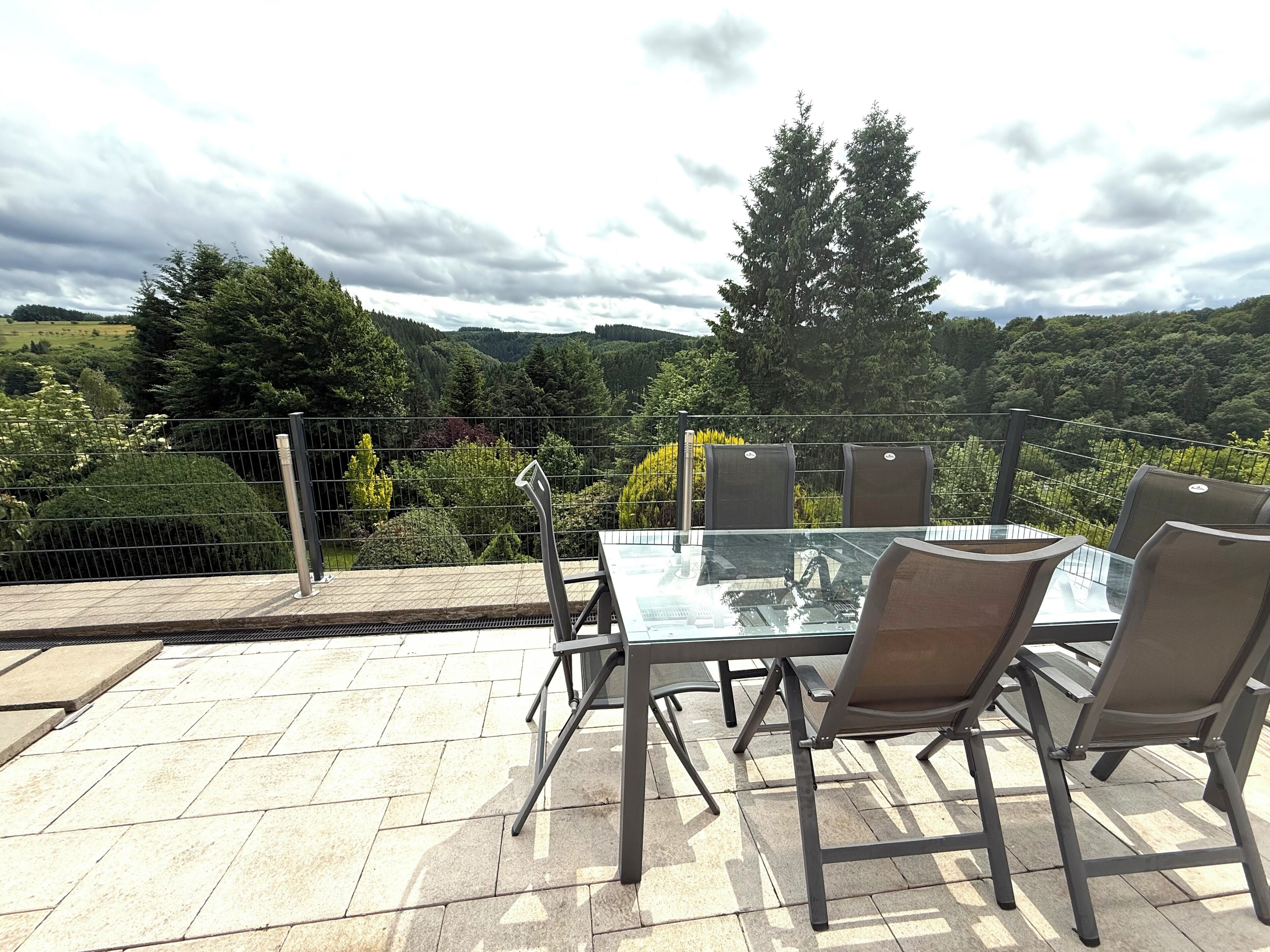 An outdoor patio with a glass table and chairs, surrounded by a metal fence, overlooking a scenic landscape with trees and hills.