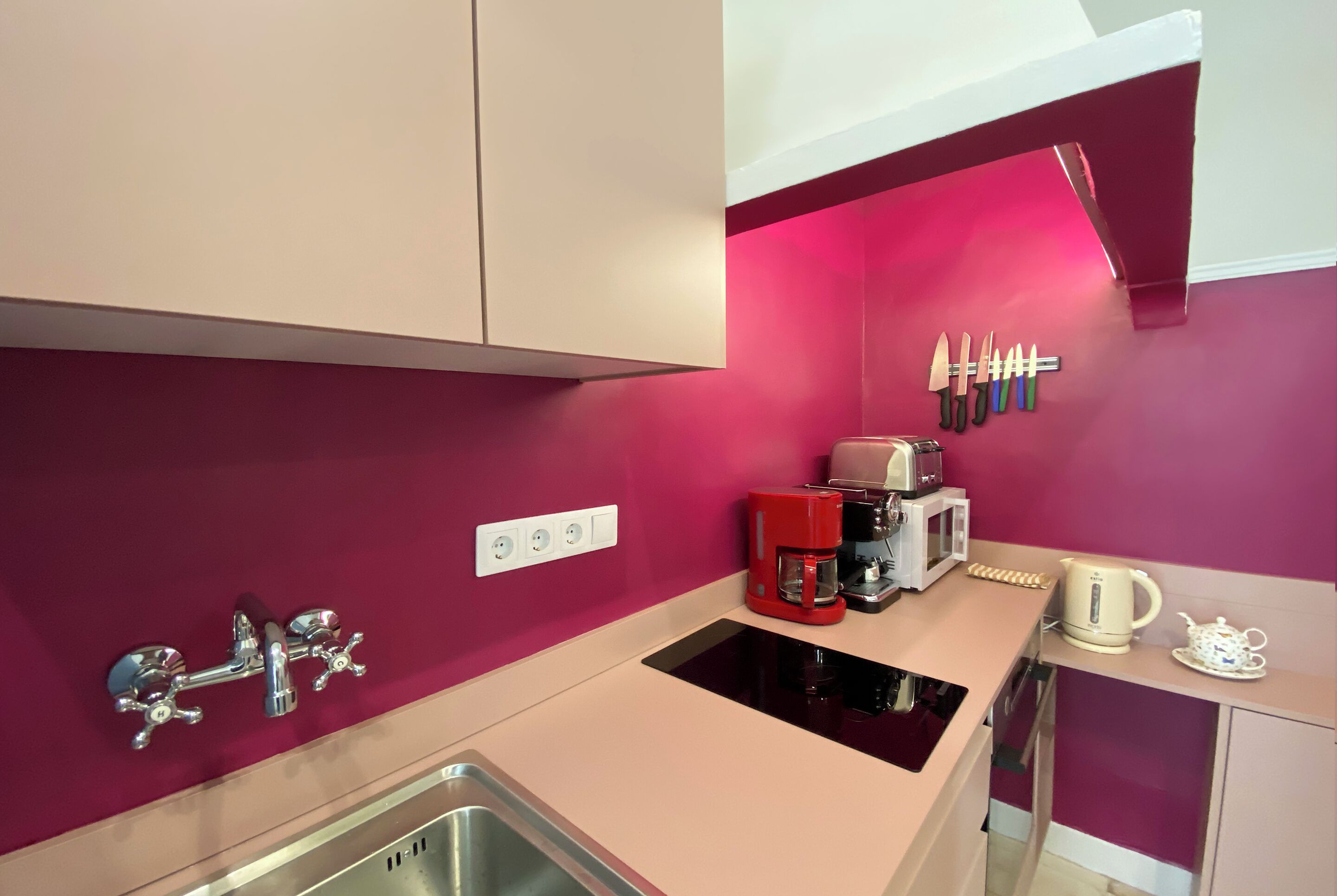 Una cocina moderna con pared rosa, pileta de acero inoxidable y cafetera roja.