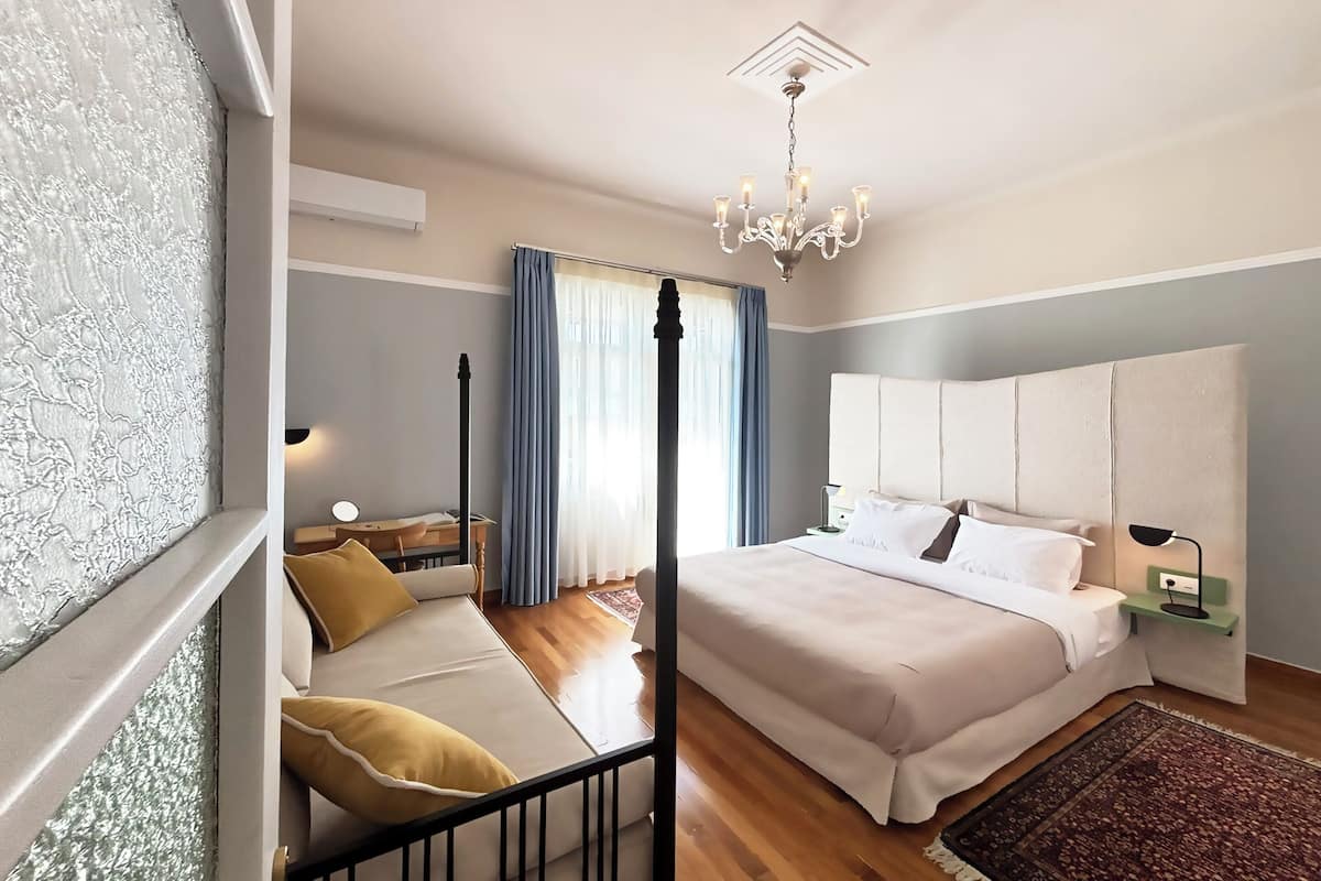 Une chambre à coucher avec un grand lit, une table de chevet, une chaise, un lustre et une fenêtre avec des rideaux.