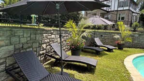 Un jardin avec un mur en pierre, une piscine et des chaises longues placées sous un grand parasol.