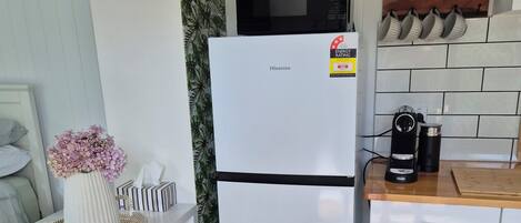Une cuisine compacte avec un réfrigérateur blanc, un four à micro-ondes et une machine à café. On y trouve une étagère avec des bocaux et un objet décoratif.