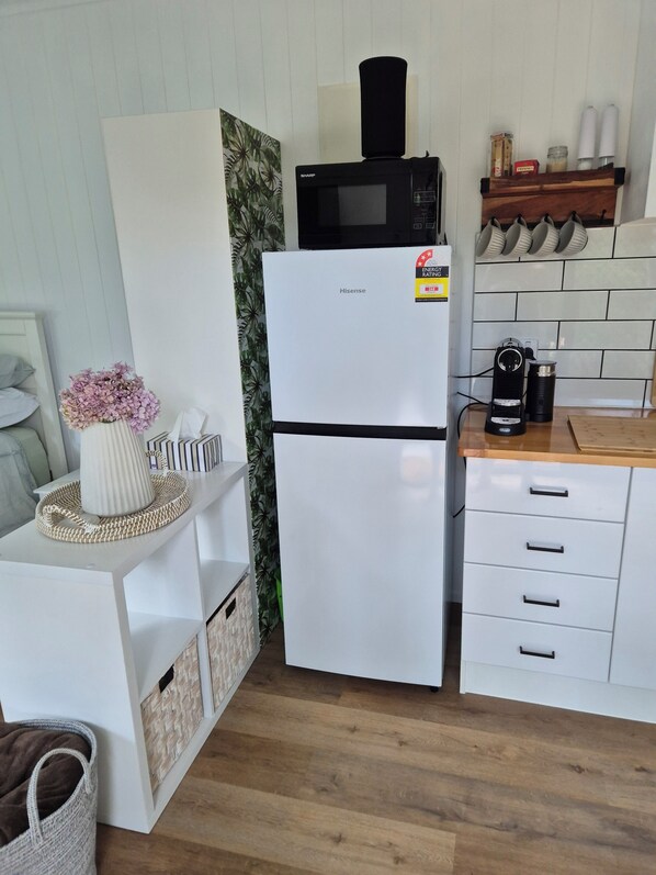 Cocina compacta con refrigerador blanco, microondas y cafetera. Hay un estante con frascos y un objeto decorativo.