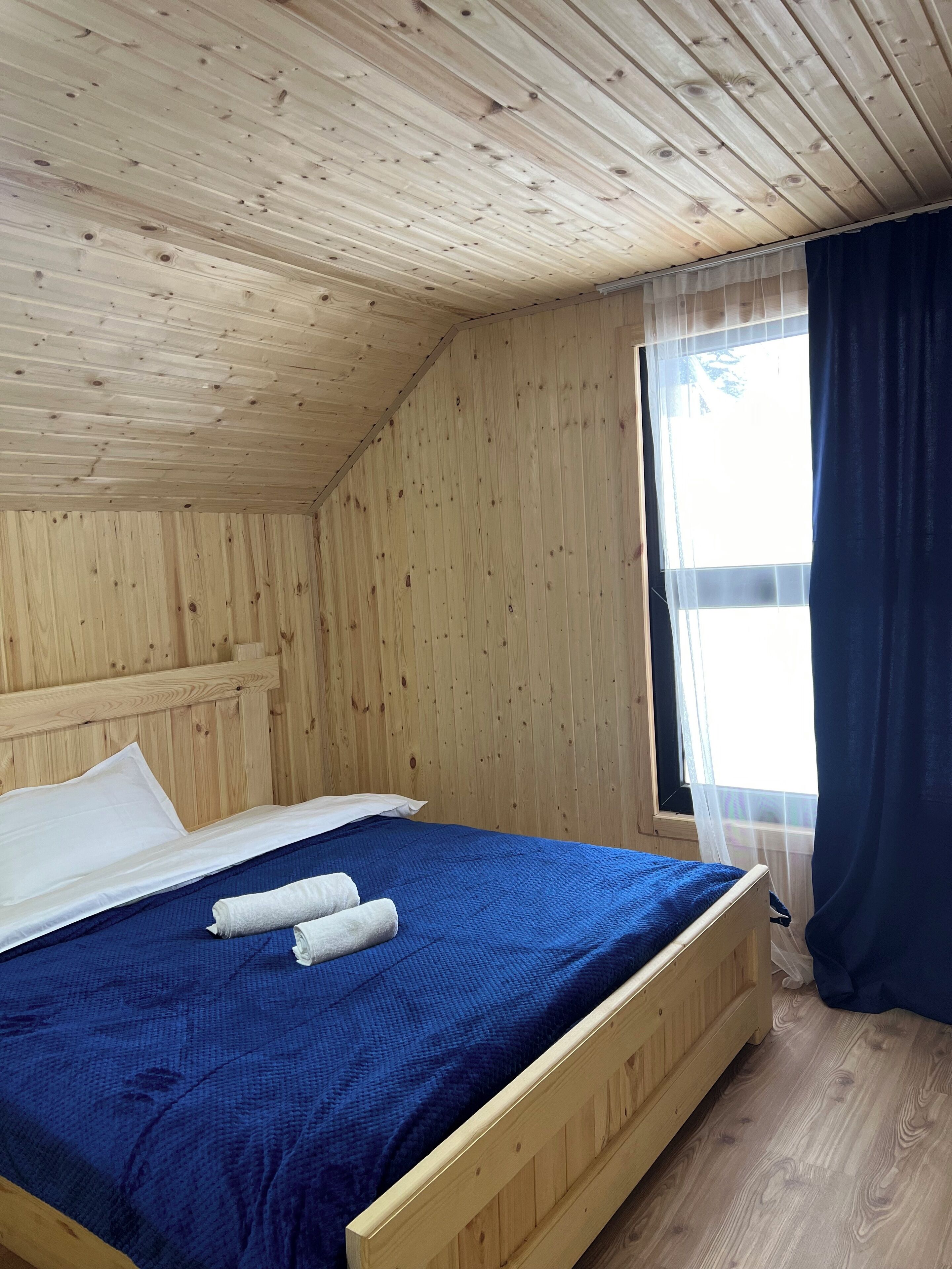 A wooden room with a bed, a window with curtains, and a wooden ceiling.