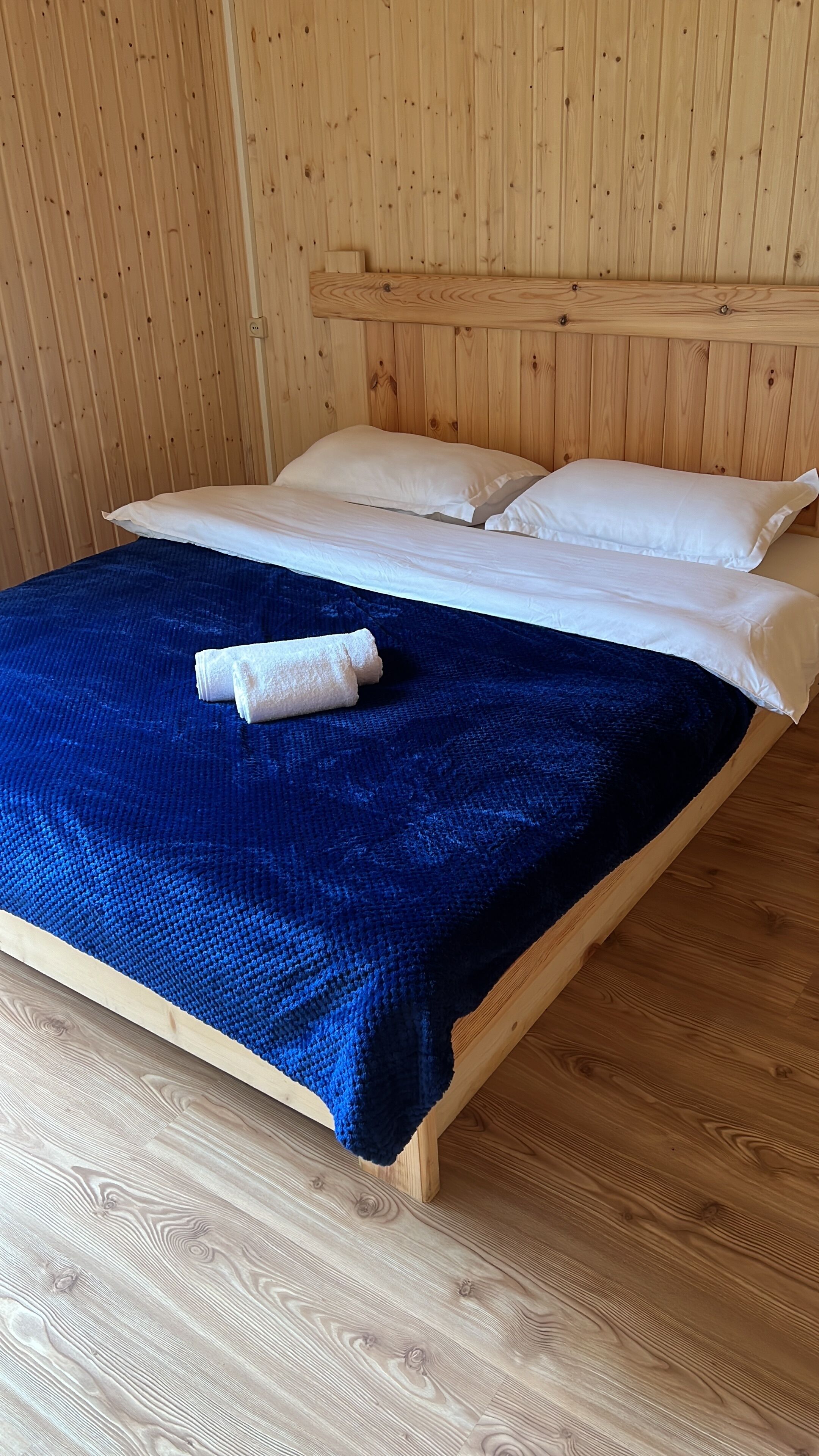 A wooden bed with white bedding and a blue blanket in a room with wooden walls and flooring.