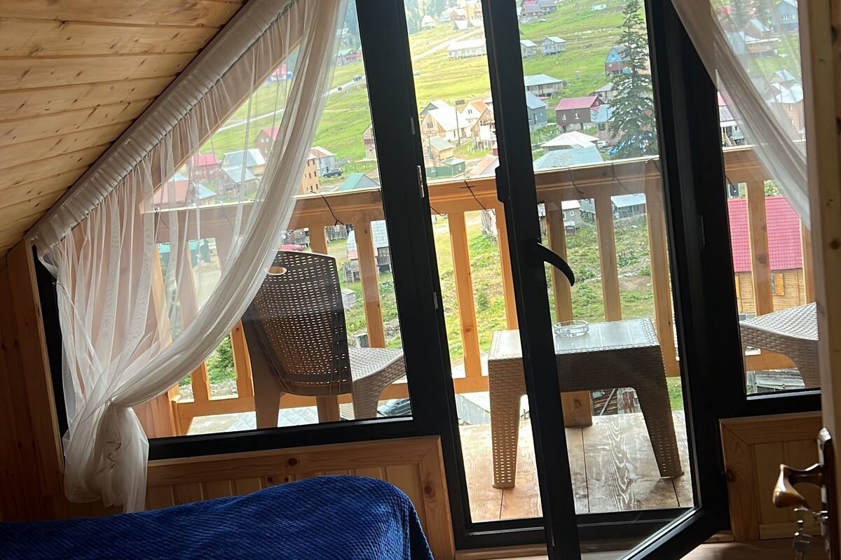 A cozy attic bedroom with a large bed, wooden ceiling, and a view of a green landscape through open doors.