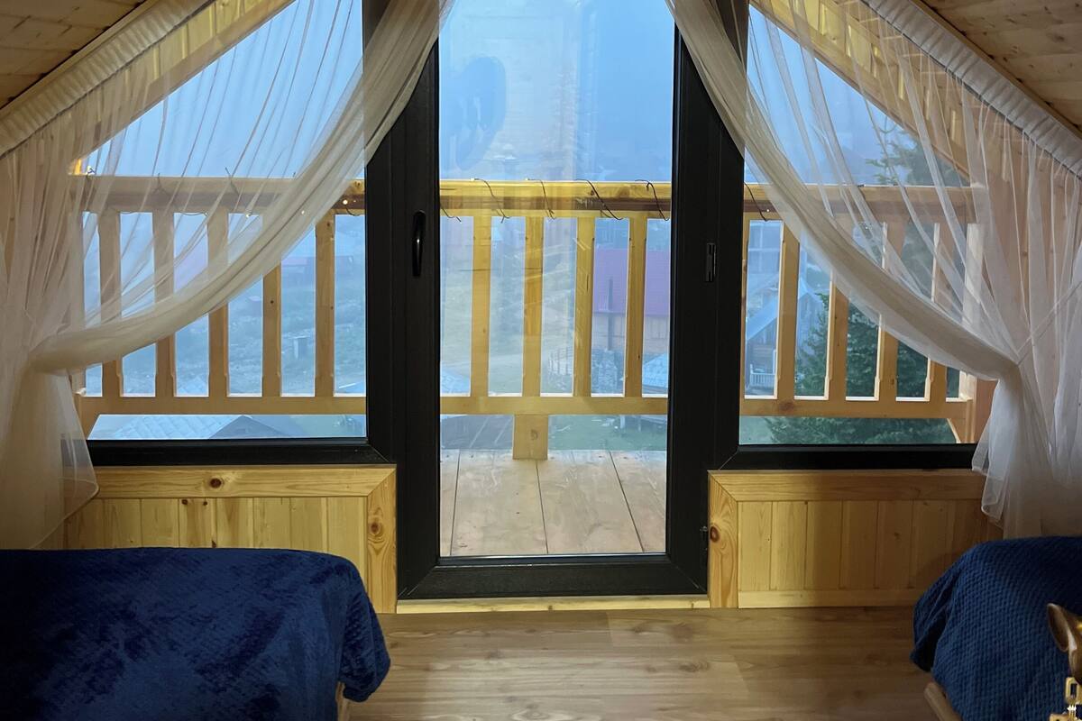 A cozy attic bedroom with a sloped ceiling, two beds, and large windows with sheer curtains.