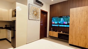 A modern bedroom with a large bed, a TV mounted on a wooden wall, and a kitchen area with cabinets and appliances. - Goba Peaks (Dar es Salaam)