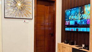 A modern living room with a large flat-screen TV displaying a streaming service interface, a wooden door, a framed abstract painting, and a built-in wooden cabinet with a phone and a glass. - Goba Peaks (Dar es Salaam)