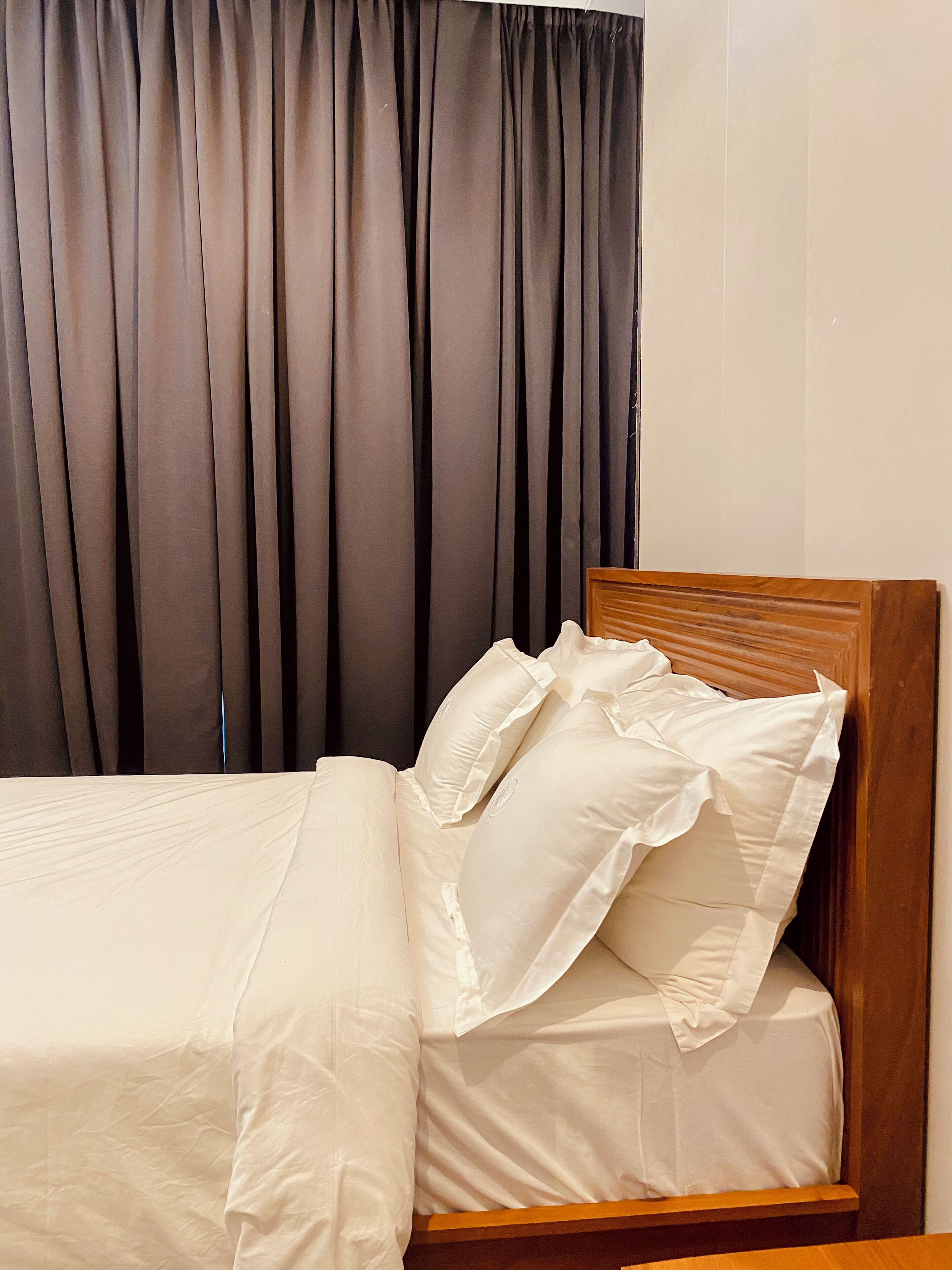 A neatly made bed with white bedding and pillows, a wooden headboard, and a dark curtain in the background.