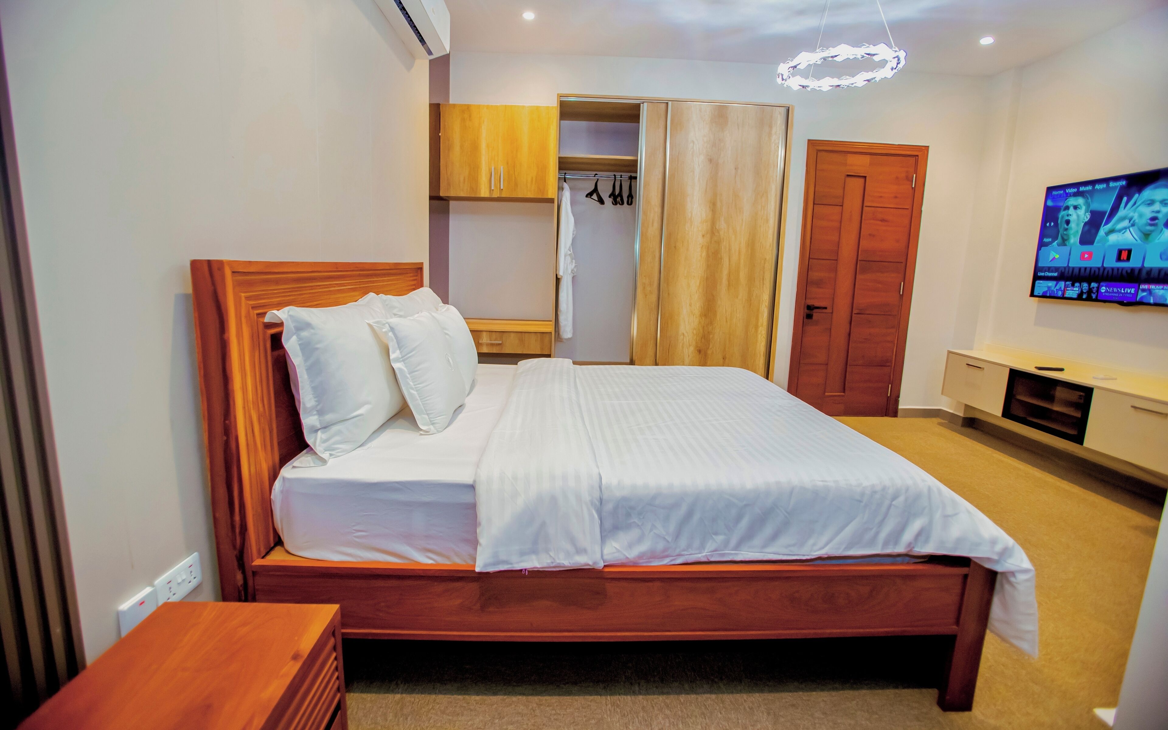 Une chambre bien rangée, avec un lit en bois, une armoire, un téléviseur et un luminaire fixé au plafond.
