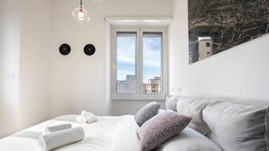 A modern bedroom with a large bed, two wall-mounted clocks, a window with a city view, and a marble accent wall.