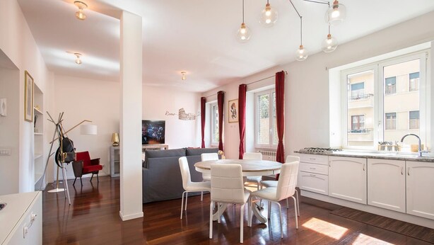 A modern kitchen with white cabinets, a dining area with a round table and white chairs, and a living area with a gray sofa.