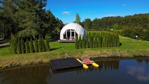 Uma tenda em forma de cúpula com um deck e área de estar, rodeada por uma floresta e um corpo d'água.