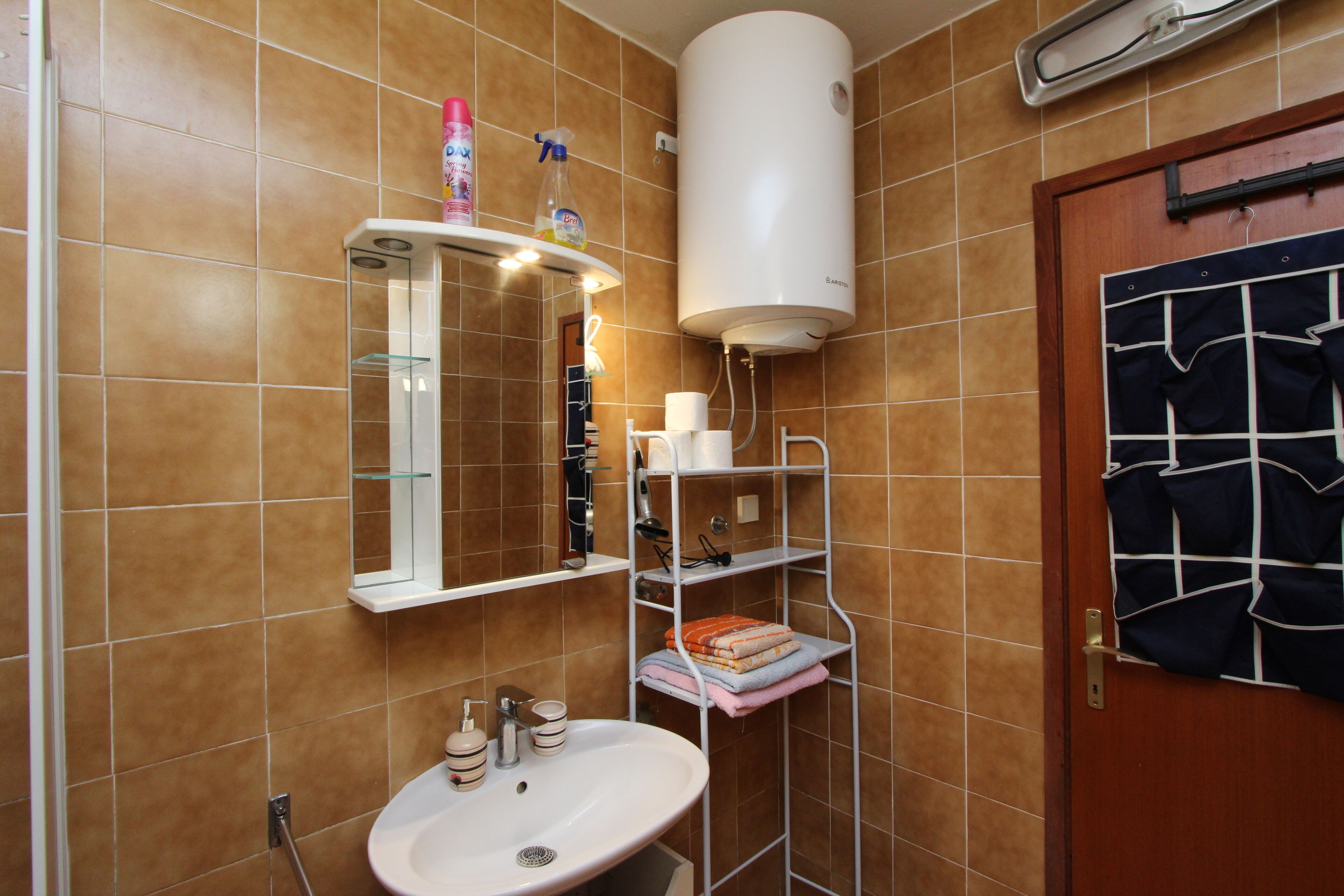 Uma casa de banho com uma pia branca, espelho e suporte para toalhas. Há um aquecedor branco na parede e uma porta com um porta-toalhas.