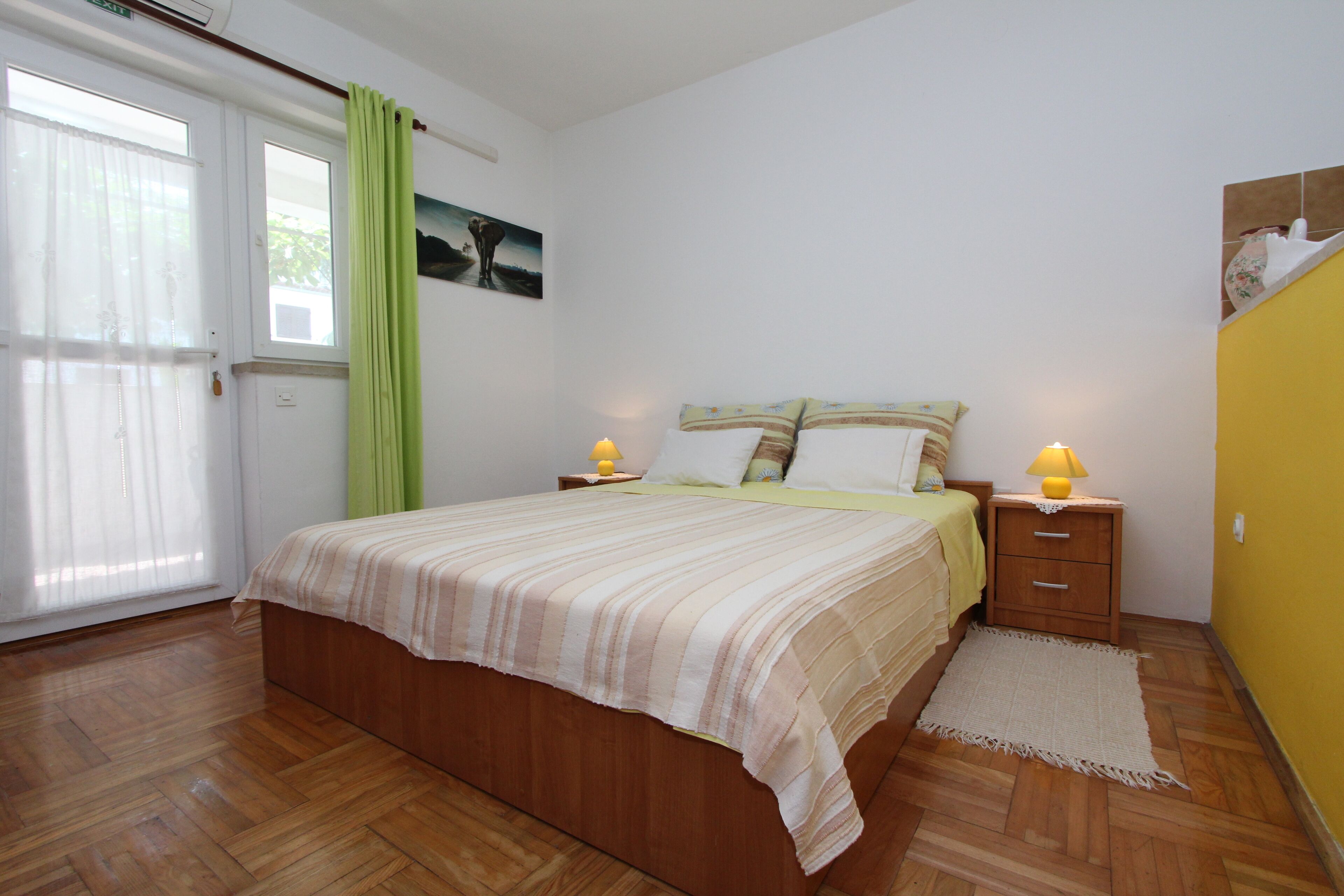 Un dormitorio con cama de madera, cortina verde, mesita de noche con lámpara y un cuadro en la pared.