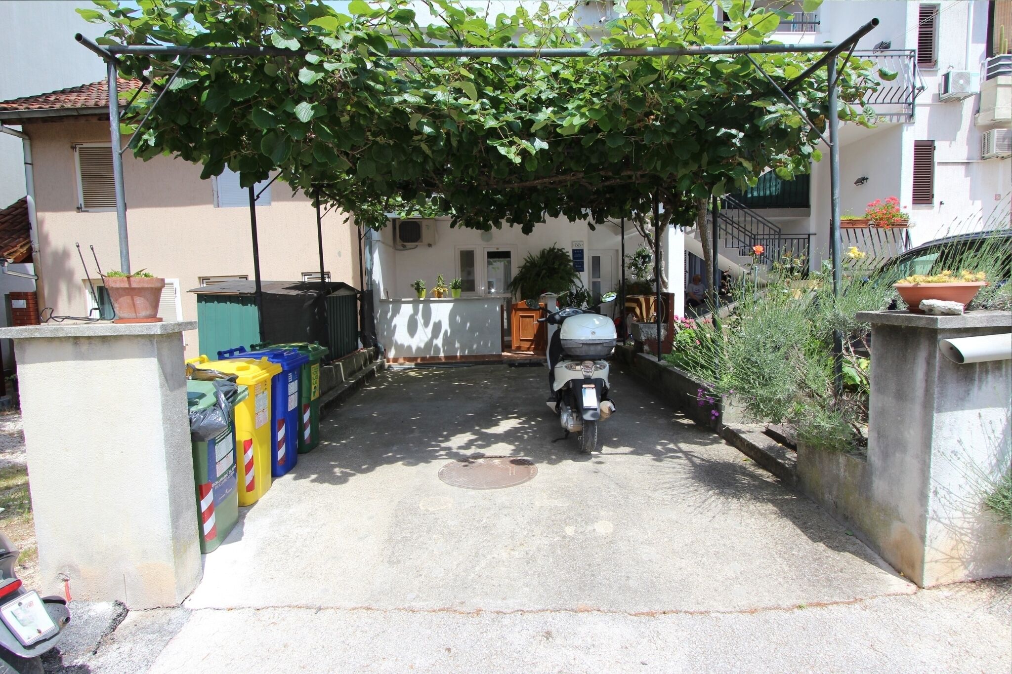 Uma entrada estreita com uma scooter estacionada e uma fileira de recipientes coloridos à esquerda.