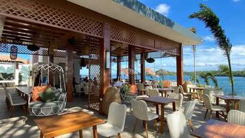 An outdoor seating area with tables and chairs, a wooden pergola, and a view of the sea.