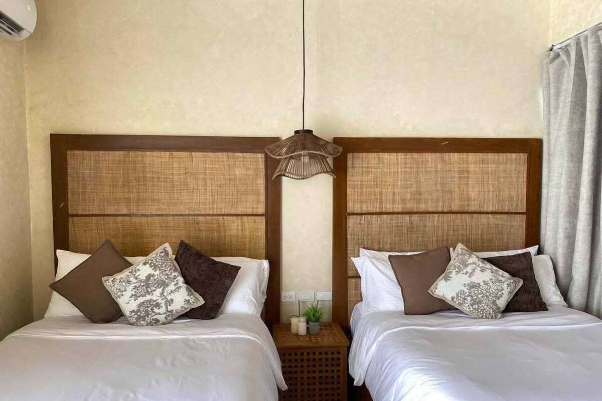Een hotelkamer met twee bedden, elk voorzien van een houten hoofdeinde en witte beddengoed. Tussen de bedden staat een klein tafeltje met een plant en een lamp.