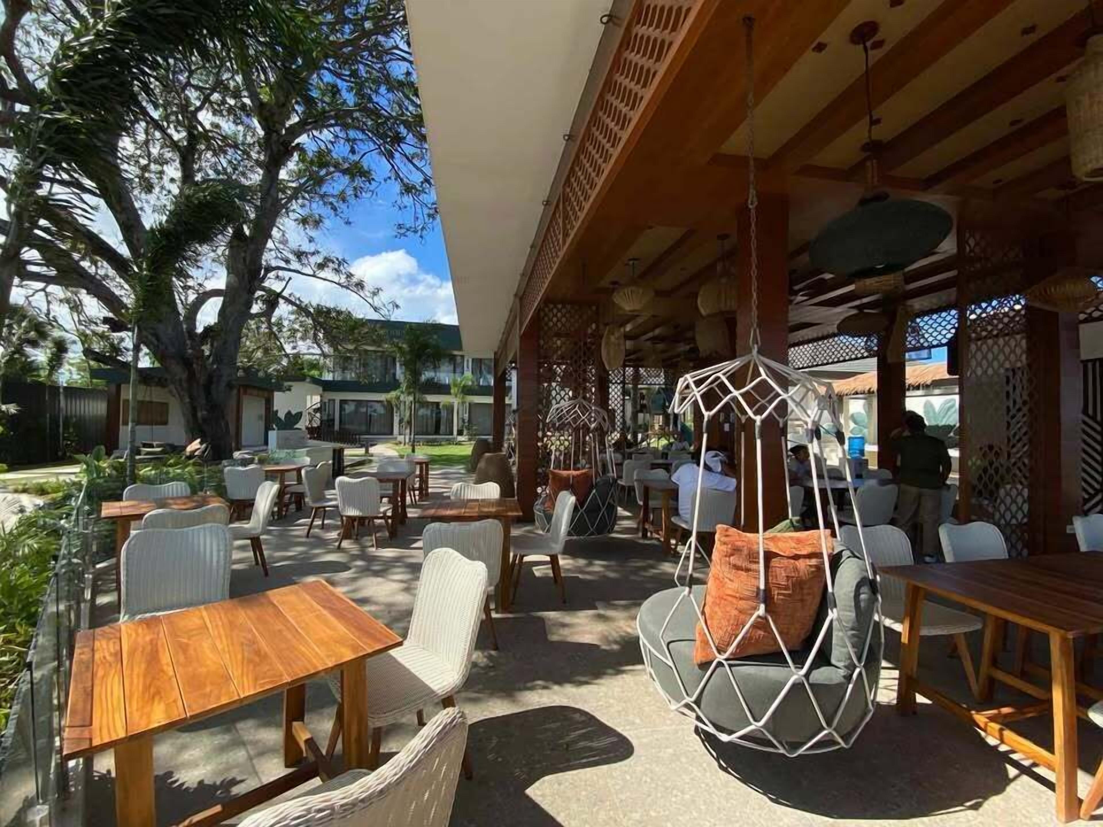 An outdoor seating area with wooden tables and chairs, a hanging chair, and a covered section with a lattice design.