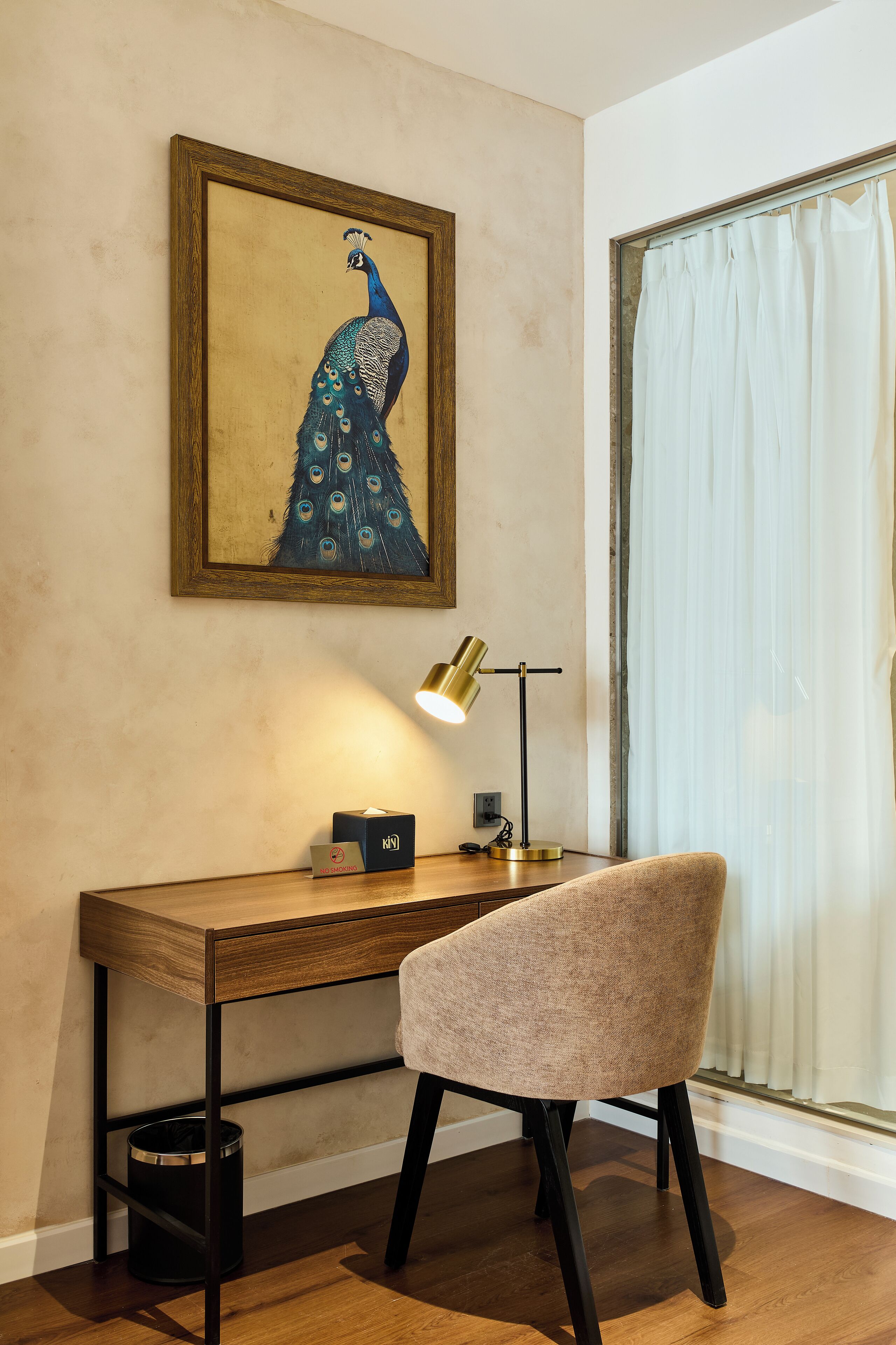 A desk with a lamp, a chair, and a framed peacock painting on the wall.
