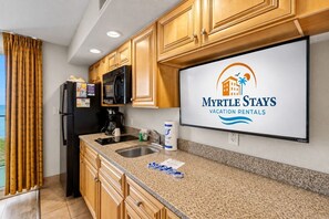 A kitchen with wooden cabinets, a black refrigerator, a microwave, and a sink. There's a large screen displaying 'Myrtle Stays Vacation Rentals' logo.