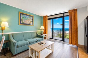 A room with a sofa, a table, a painting, and a view of the beach.