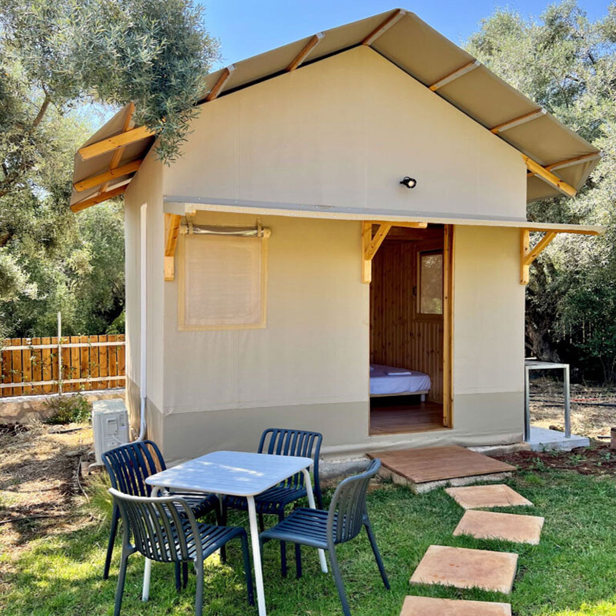 A small, single-room cabin with a bed, a table, and chairs in the yard.
