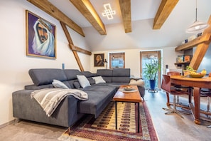 A modern living room with a grey sofa, wooden beams, a dining table with chairs, and a large window.