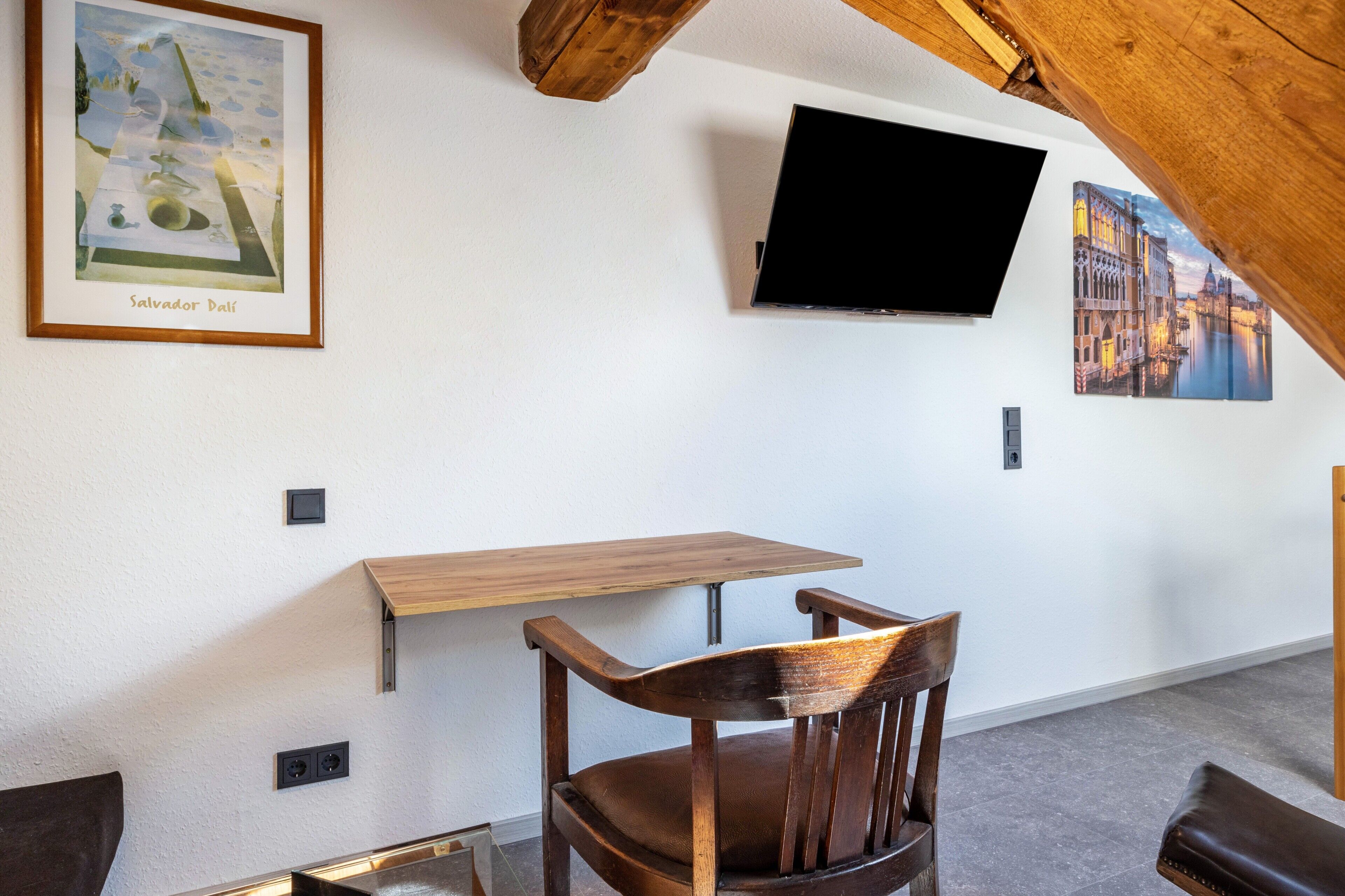 Une pièce avec un plafond en bois, un téléviseur fixé au mur, un tableau encadré représentant Salvador Dali et une table en bois avec une chaise.