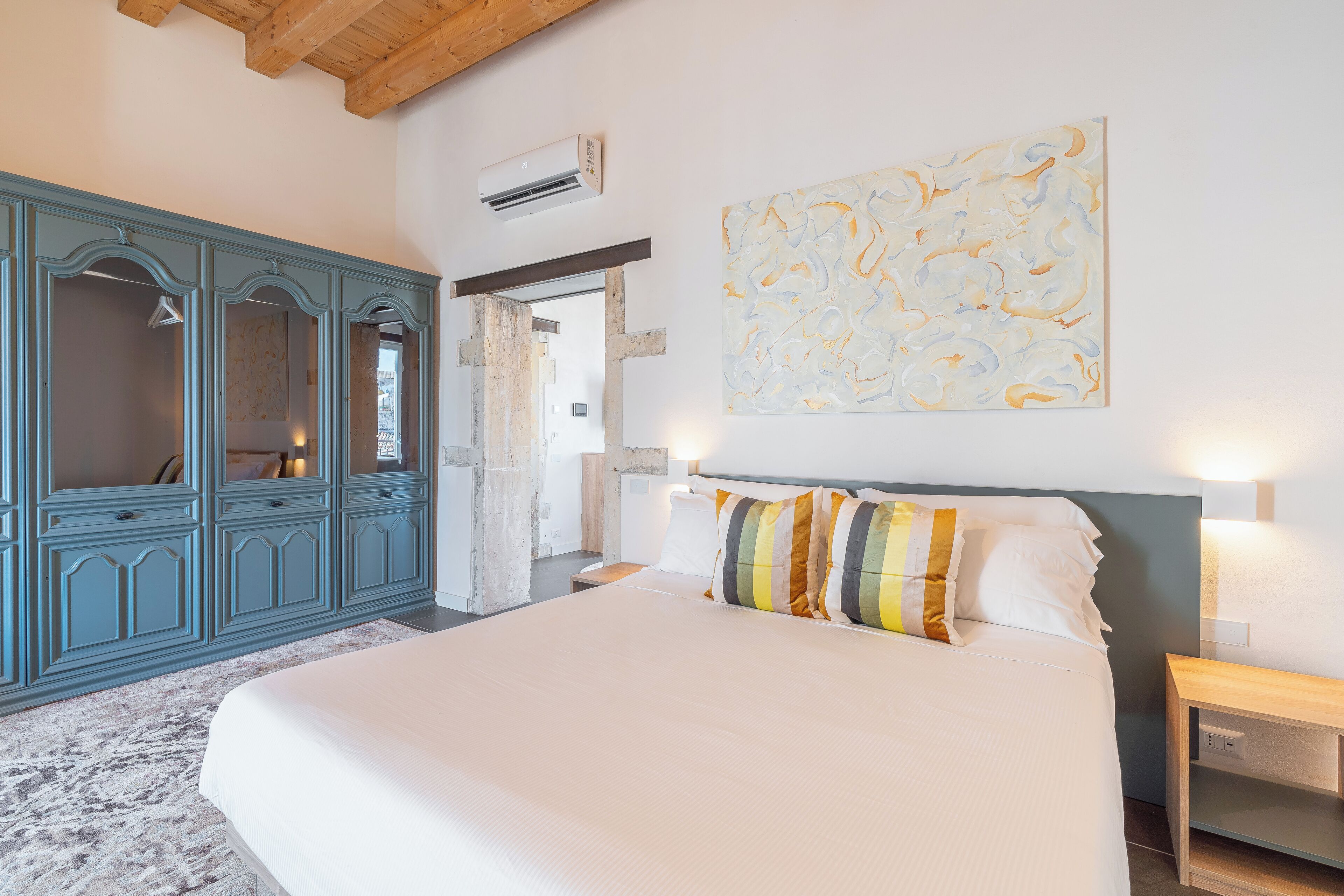 A bedroom with a large bed, a blue wardrobe, a painting on the wall, and an air conditioning unit.