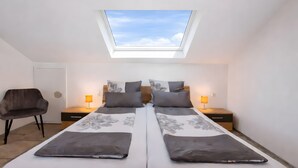 A bedroom with a skylight, two beds with grey floral bedding, bedside lamps, a chair, and a wooden nightstand. - Casa di Napolitano at Lake Constance – Modern Attic Apartment Near the Lake (Radolfzell am Bodensee)