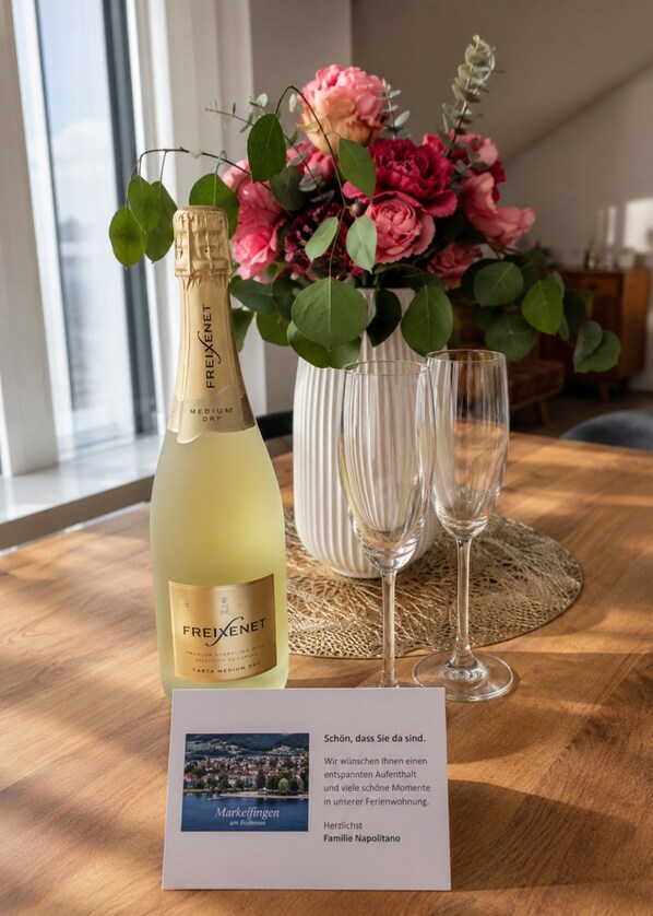 Una botella de vino Freixenet, dos copas, un ramo de flores y una tarjeta de bienvenida sobre una mesa de madera.