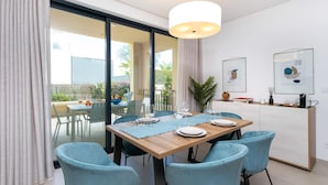 A dining area with a wooden table set for four, surrounded by teal chairs, a glass partition, and a view of an outdoor patio. - Terrazza Beach Golf (Loulé)