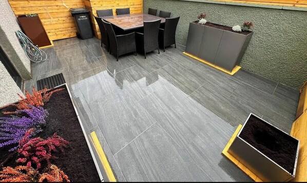 Un patio con una mesa y sillas para comer, una maceta con plantas y un basurero.