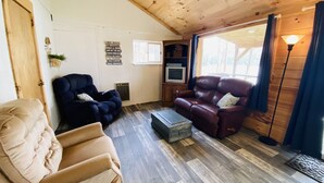 Un salon chaleureux avec un plafond en bois, un fauteuil beige, un fauteuil bleu foncé, un canapé en cuir rouge et un petit téléviseur posé sur un meuble en bois.