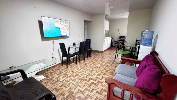 A living room with a TV mounted on the wall, a dining area with chairs and a table, a water dispenser, and a sofa with a cushion. - Peace N' Green (David)
