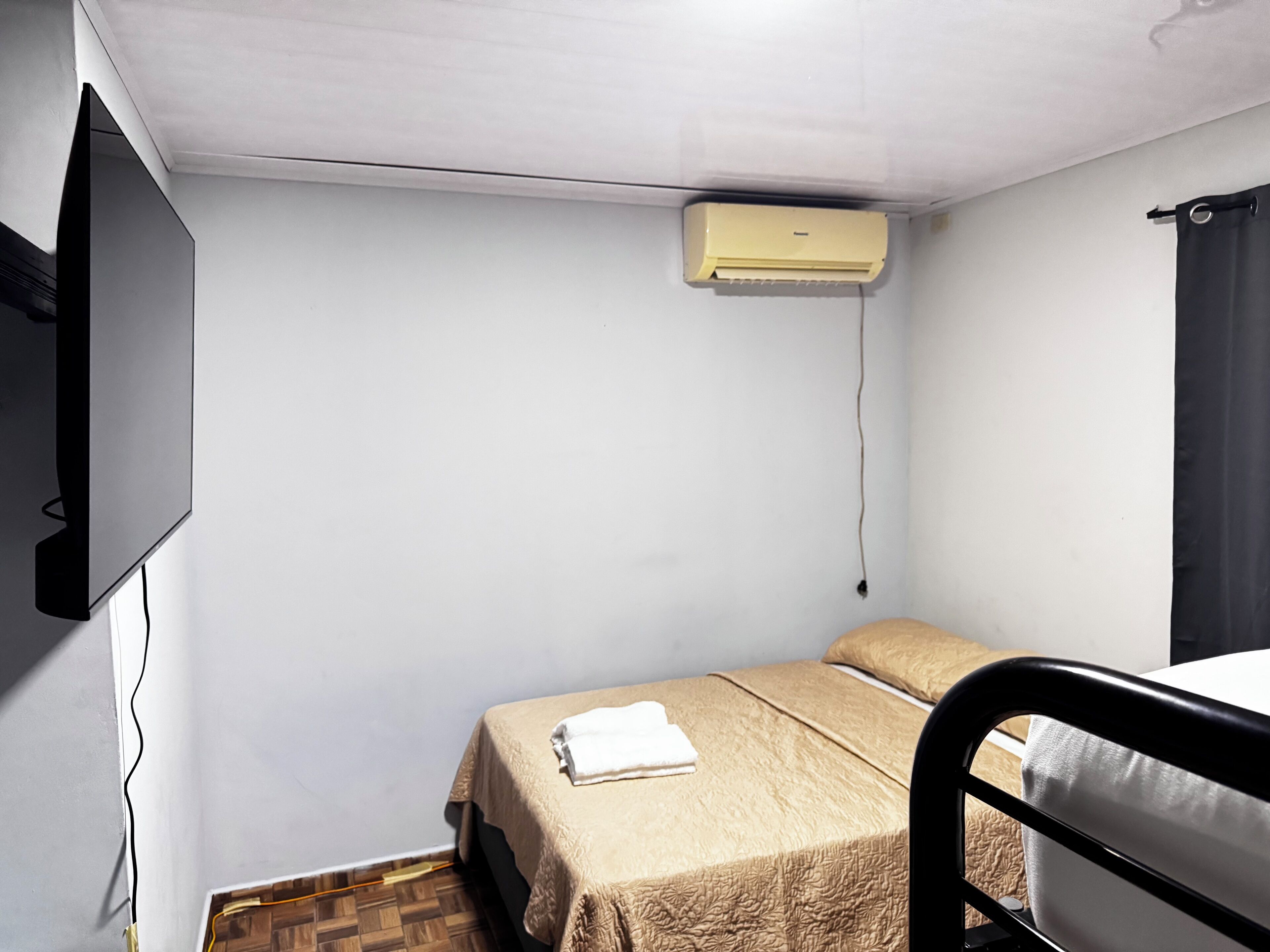Quarto pequeno com uma cama de solteiro, televisão fixada na parede, ar-condicionado no teto, manta bege e toalha branca.